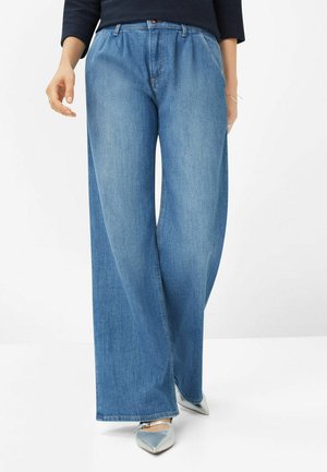 Personne portant un jean bleu à jambes larges et des chaussures argentées à bouts pointus, debout devant un fond blanc uni.