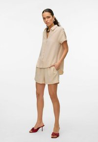 Beżowa koszula z krótkim rękawem, zestawiona z pasującymi szortami. Modelka ma na sobie czerwone sandały na obcasie z kwiatowymi zdobieniami.