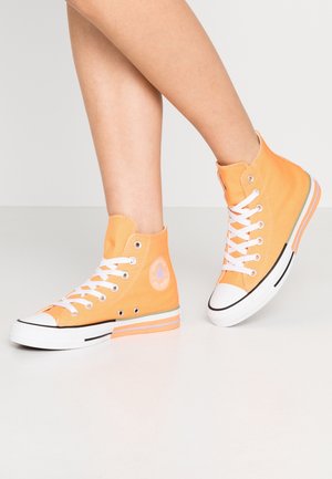 Henkilö, jolla on oranssit korkeavartiset Converse-tennarit, joissa on valkoiset nauhat ja pohjat, seisoo vaalealla yksivärisellä lattialla.