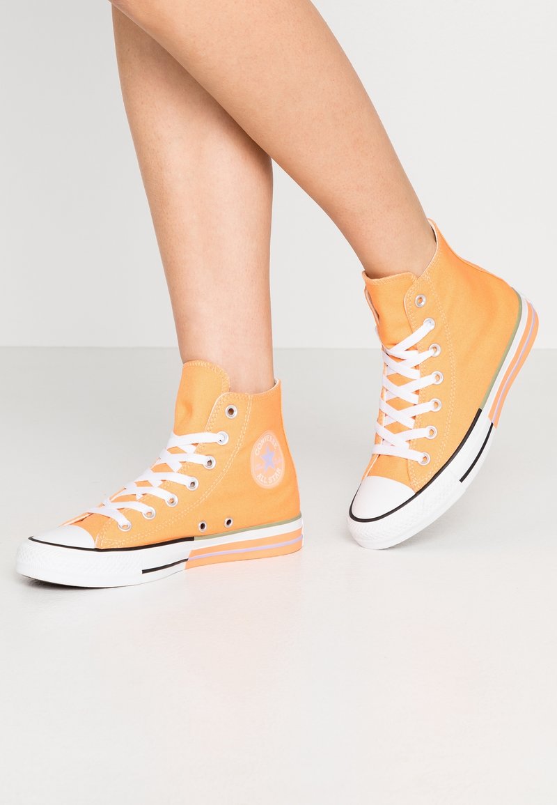 Personne portant des baskets montantes Converse orange avec des lacets et des semelles blanches, debout sur un sol uni et clair.