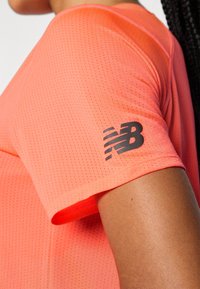 Trumpa rankovėmis sportinė maikutė ryškiai koralų spalvos, pagaminta iš lengvos, kvėpuojančios medžiagos su tekstūruotu raštu ir juodu logotipu ant rankovės.