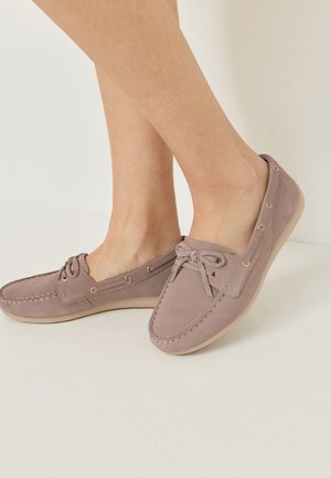 Nohy v mauve semišových mokasínach s čipkovými detailmi a béžovými gumovými podrážkami na svetlej podlahe a stene.
