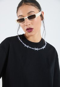 Camiseta de algodón negra con bordado de alambre de púa blanco en el escote. Acompañada de gafas de sol rectangulares color crema.