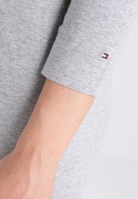 Tissu gris avec une texture douce, présentant une manche retroussée. Comprend une petite étiquette de logo en bleu, rouge et blanc au poignet.