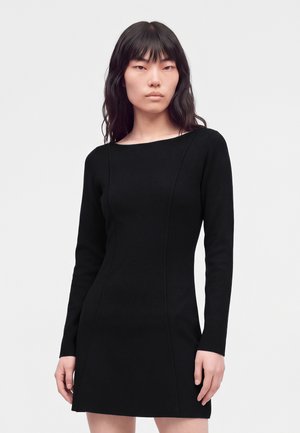 Femme aux longs cheveux foncés portant une robe noire ajustée à manches longues, debout devant un fond clair uni.
