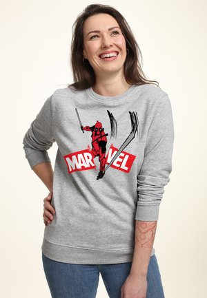 Uśmiechnięta kobieta nosząca szarą bluzę Marvel z postacią Deadpoola z mieczami w pozie akcji na przodzie, stojąca z ręką na biodrze.