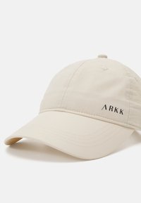 Beige kerge kork, millel on kaardus visor, esipaneelil mustalt brodeeritud logo "ARKK" ja peal väiksed ventilatsiooniavad.