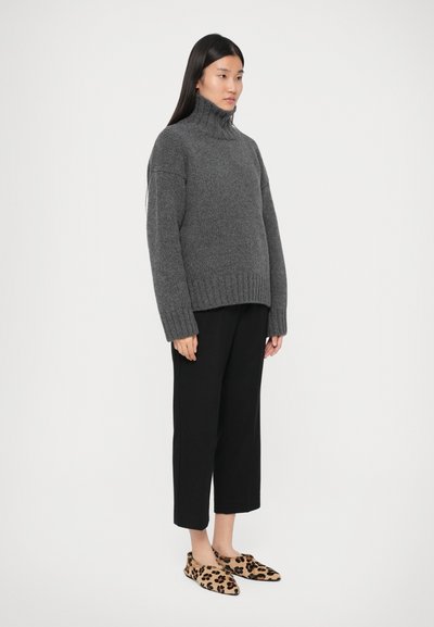 Donna che indossa un maglione grigio a collo alto, pantaloni neri a culotte e scarpe slip-on con stampa leopardata, in piedi contro uno sfondo bianco uniforme.