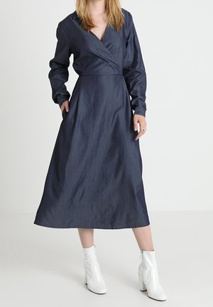 Langärmliges Wickelkleid aus dunkelblauem Denim mit figurnahem Oberteil und weit ausgestellt Rock, mit seitlichen Taschen und glatter Textur. Kombiniert mit weißen Stiefeln.