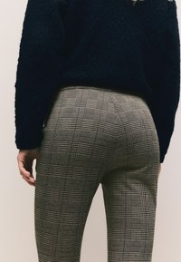 Person iført sort tekstureret sweater og tætsiddende grårute bukser vist fra midten af ryggen til midten af låret, stående med venstre hånd afslappet.