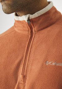 Pomarańczowy polarowy pulower z wysokim kołnierzem i półzamkiem. Posiada kontrastowy biały wnętrze kołnierza oraz subtelne logo marki.