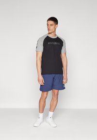 Svart og grå T-skjorte med kort erm og "HUMMEL"-logo, kombinert med blå shorts. Modellen har på seg hvite sneakers og hvite sokker. Enfarget bakgrunn.