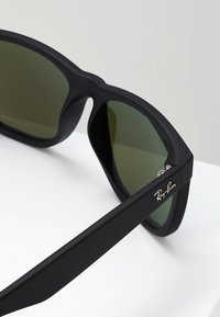 Ochelari de soare negri mat cu lentile verzi, având un design geometric elegant și un logo Ray-Ban argintiu pe lateral.