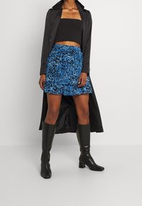 Femme portant un crop top noir et un long manteau noir avec une jupe à imprimé léopard bleu et noir et des bottes à talons hautes noires jusqu'aux genoux.