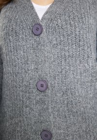 Cardigan grigio lavorato a maglia con scollo a V, tre grandi bottoni viola e un tessuto texturizzato con costine verticali.
