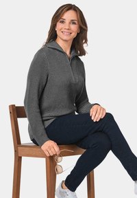 Grijze gebreide trui met een kwart-rits kraag, gecombineerd met donkere jeans en witte sneakers. Accessoires zijn een bril die in de hand wordt gehouden.