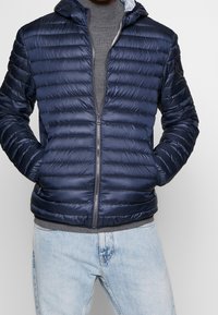 Marinblå puffjacka med horisontellt kviltad textur, full dragkedja och en huva. Bärs över en grå stickad tröja och ljusblå jeans.