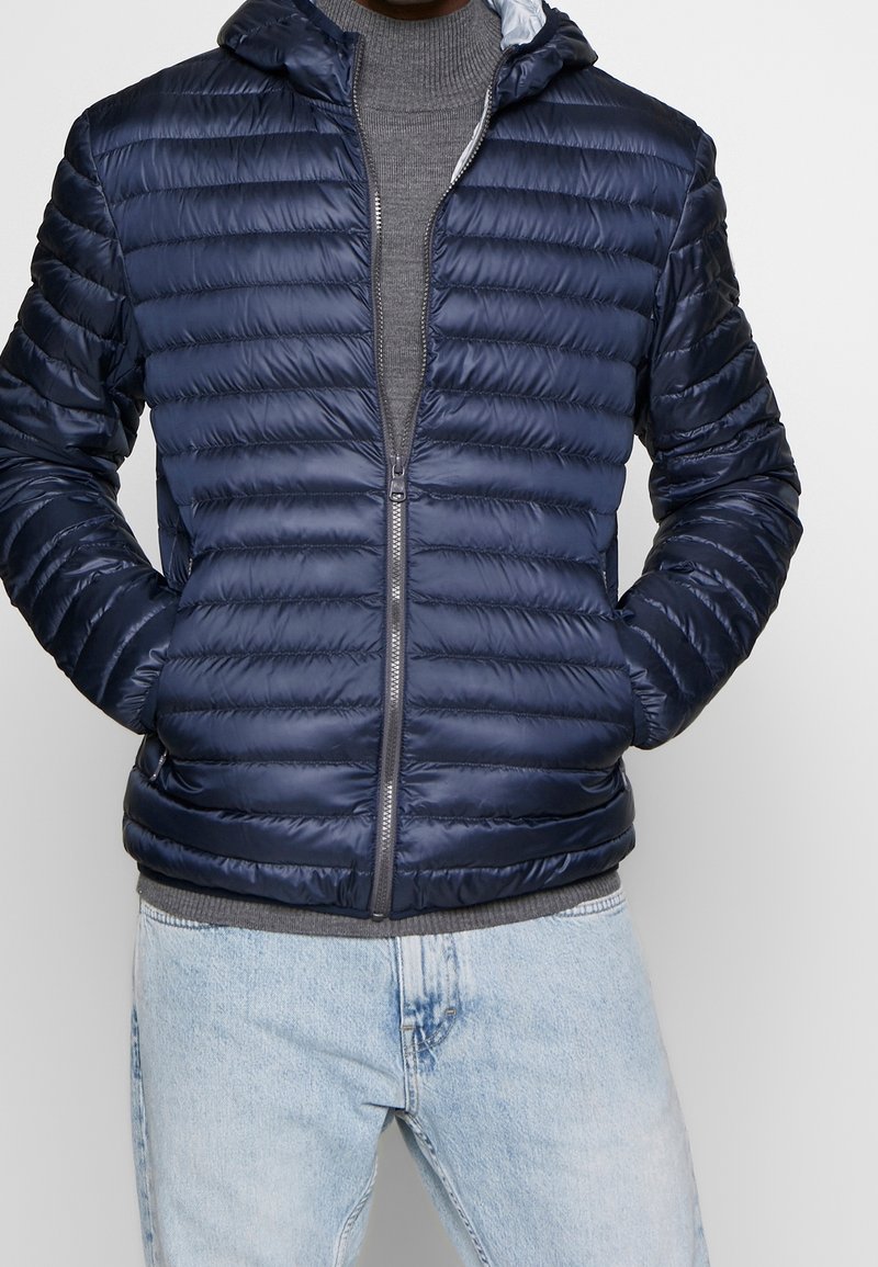 Marinblå puffjacka med horisontellt kviltad textur, full dragkedja och en huva. Bärs över en grå stickad tröja och ljusblå jeans.