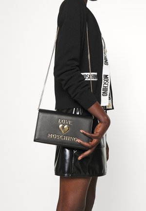 Czarna skórzana torebka typu crossbody z złotym napisem "LOVE" i detalem w kształcie serca. Gładka tekstura, prostokątny kształt i łańcuszkowy pasek. Stylizowana z czarną bluzką.