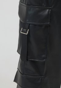 Schwarzer Kunstlederrock-Detail mit einer Klappentasche, die durch einen schwarzen Riemen und eine silberne rechteckige Schnalle gesichert ist.