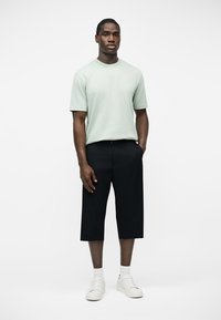 Άνδρας που στέκεται, φοράει ανοιχτό πράσινο κοντομάνικο πουκάμισο, μαύρο φαρδύ παντελόνι μέχρι τον αστράγαλο, λευκές κάλτσες και λευκά αθλητικά παπούτσια, σε λευκό φόντο.