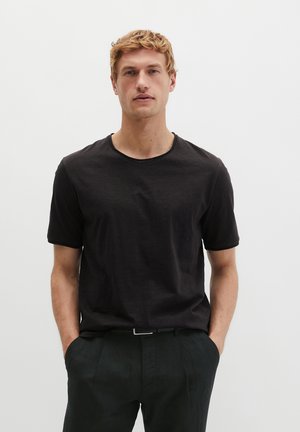 Man met licht krullend haar, gekleed in een effen zwart T-shirt met korte mouwen en een zwarte broek, staand met handen in de zakken tegen een witte achtergrond.
