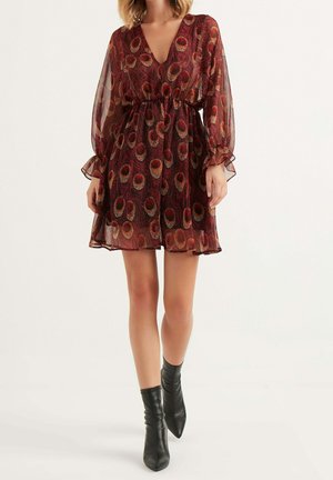 Nainen pukeutuneena läpikuultavaan pitkähihaiseen minimekkoon, jossa on punainen ja kultainen riikinkukon sulkakuvio, sekä mustat nilkkurit, seisomassa yksinkertaisen valkoisen taustan edessä.