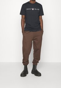 Camiseta navy con el logo "GANT 1949", pantalones deportivos marrones y botas negras de tobillo. Presenta un corte clásico y puños acanalados en los pantalones.