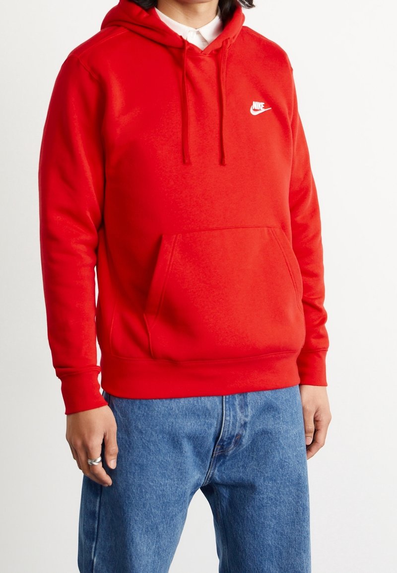 Personne portant un sweat à capuche Nike rouge vif avec poche avant et cordons de serrage, associée à un jean bleu sur un fond blanc uni.