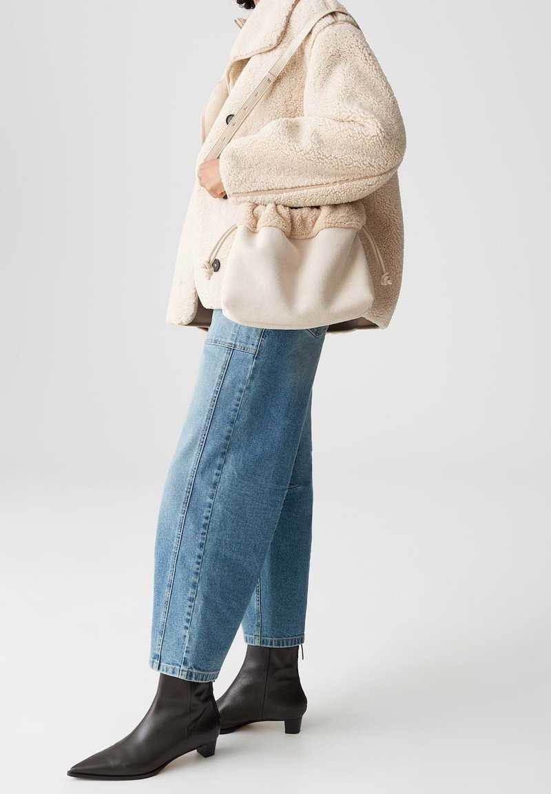 Personne portant un manteau en peluche beige clair, un jean en denim bleu clair, des bottines noires et tenant un sac à main beige doux avec une bandoulière.
