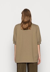 Khaki oversized mikina s krátkými nabíranými rukávy, vyrobená z měkkého materiálu; kombinováno s hnědými skládacími kalhotami, pohled zezadu.
