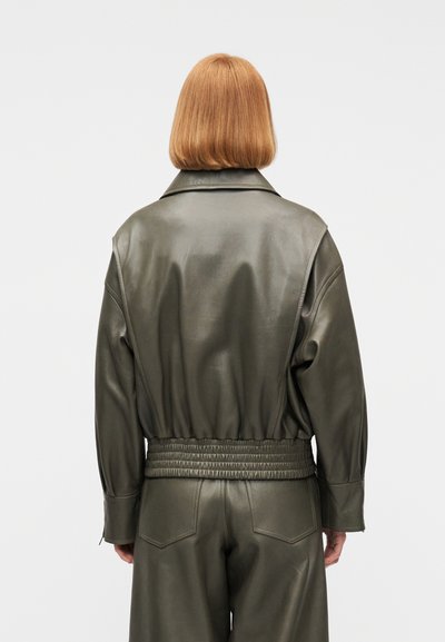 Άτομο με ίσια, κόκκινα μαλλιά μήκους έως τους ώμους, φορώντας ένα σκούρο πράσινο δερμάτινο μπουφάν και αντίστοιχο δερμάτινο παντελόνι, εμφανιζόμενο από την πίσω πλευρά.