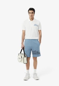 Camisa polo blanca con logo azul, pantalones cortos azul claro con un diseño impreso en negro y una bolsa blanca de transporte con acentos en negro.