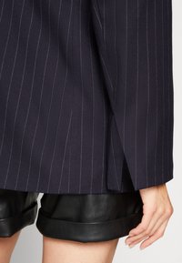 Merivoimien piikkiraitatakki pystysuorilla valkoisilla raidoilla, jossa on terävät kaulukset ja sivusaumat, yhdistetty mustiin nahkashortseihin, joissa on käännetyt lahkeet.