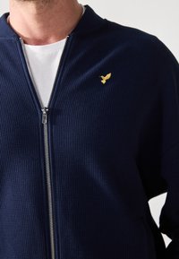Chaqueta de cremallera azul marino hecha de tejido texturizado, con un logo de pájaro dorado en el lado izquierdo del pecho y una camiseta blanca debajo.