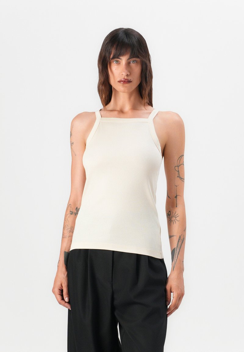 Crème ribbel tanktop met brede banden en een vierkante halslijn, gemaakt van rekbare stof; gecombineerd met loszittende zwarte broek.