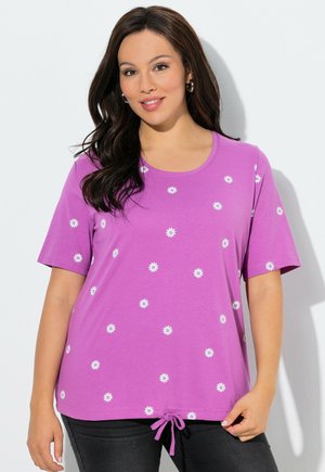 Kobieta o długich ciemnych włosach nosi fioletową koszulkę z krótkim rękawem z białymi wzorami stokrotek i ściągaczem u dołu, w połączeniu z czarnymi spodniami.