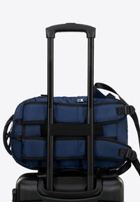 Tmavě modrá polstrovaná taška připevněná na černém kolečkovém kufru se dvěma teleskopickými rukojeťmi, vybavená nastavitelnými popruhy a zipem.