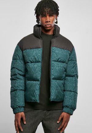 Teal pufferjack met een ingewikkeld patroon, zwarte gewatteerde schouders, hoge kraag en ritsluiting. Draagt het over een zwart shirt met grijze jeans.