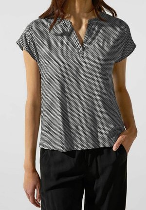 Vrouw draagt een zwart-wit gebloemde korte mouwen blouse met een V-hals en zwarte broek, één hand in de zak.