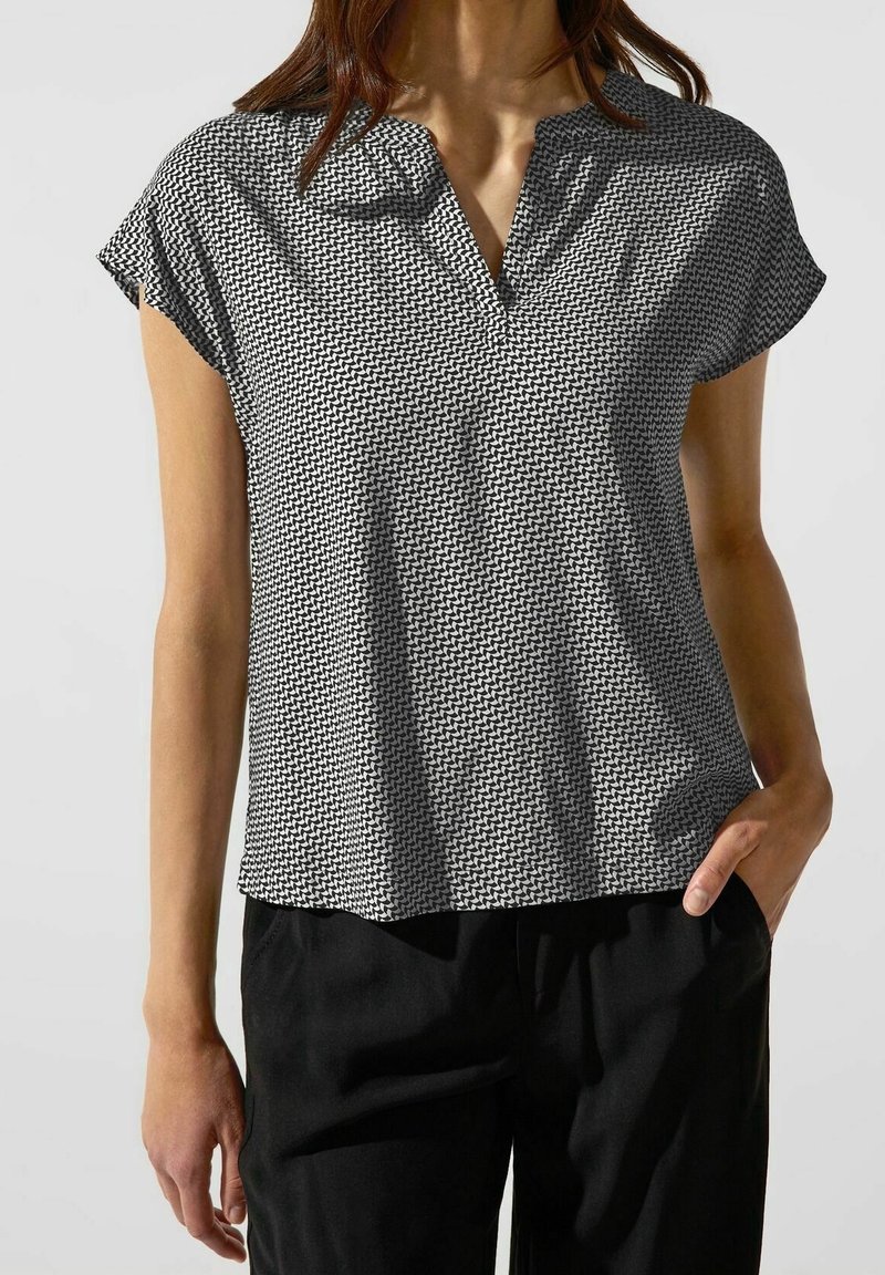Femme portant un chemisier à manches courtes noir et blanc à motifs avec un col en V et un pantalon noir, une main dans la poche.