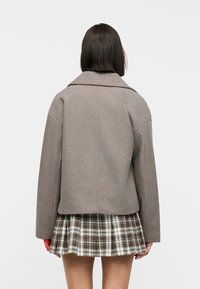 Γυναίκα με σκούρα μαλλιά μέχρι τους ώμους, φοράει ένα ραφτό μπουφάν ταμπά και μια καφέ και λευκή καρό πλισέ φούστα, στέκεται γυρισμένη με την πλάτη.