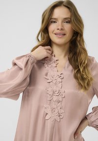 Blusa rosa empolvado de mangas largas, con detalles de encaje floral texturizado a lo largo del escote y un brillo sutil en la tela.