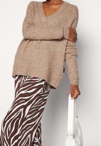 Hellbraunes V-Ausschnitt-Pullover aus weichem Material; kombiniert mit weiß-braun gestreiften Zebra-Hosen; hält eine weiße Handtasche.