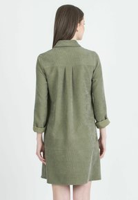 Abito in corduroy verde oliva con maniche lunghe, pieghe sulle spalle e polsini arrotolati. Texture liscia, lunghezza fino a metà coscia e design semplice.
