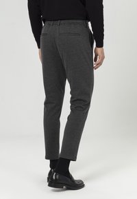 Graue Hose mit schmaler Passform, elastischem Bund und zwei Gesäßtaschen; kombiniert mit schwarzen Schuhen und dunklen Socken. Glatte Textur.