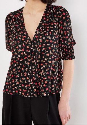 Blouse noire avec petit imprimé floral rouge et rose, col en V, manches courtes bouffantes avec poignets smockés, portée avec un pantalon noir.