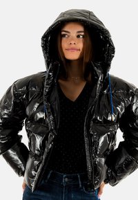 Giacca a vento nera con finitura lucida, dotata di cappuccio, stringhe blu, chiusura con zip frontale e design trapuntato. Indossata sopra una camicia nera a pois.