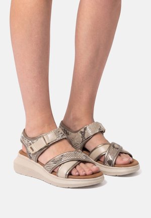 Pieds portant des sandales beiges à motif serpent avec des sangles ajustables et des semelles beiges rembourrées, posés sur un fond uni gris clair.