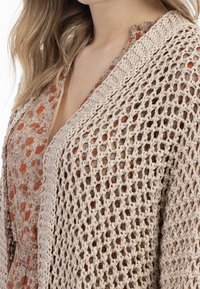 Beżowy sweter robiony na drutach o otwartym splocie, z teksturowaną powierzchnią. W tle znajduje się odzież w kwiatowy wzór w ciepłych odcieniach.